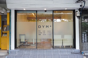 DYM+クリニックの正面玄関。ガラスの自動ドアにはロゴと日本語で書かれた診療時間が表示されている。