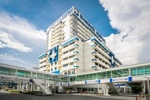 青い文字で「BANGKOK HOSPITAL」と書かれた、白と水色の高層病院の外観。手前には複数の連絡通路があり、青空と白い雲が背景に見える。