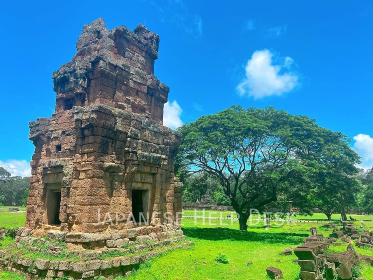 青空の下、緑の草原に立つ石造りの遺跡と大きな木。